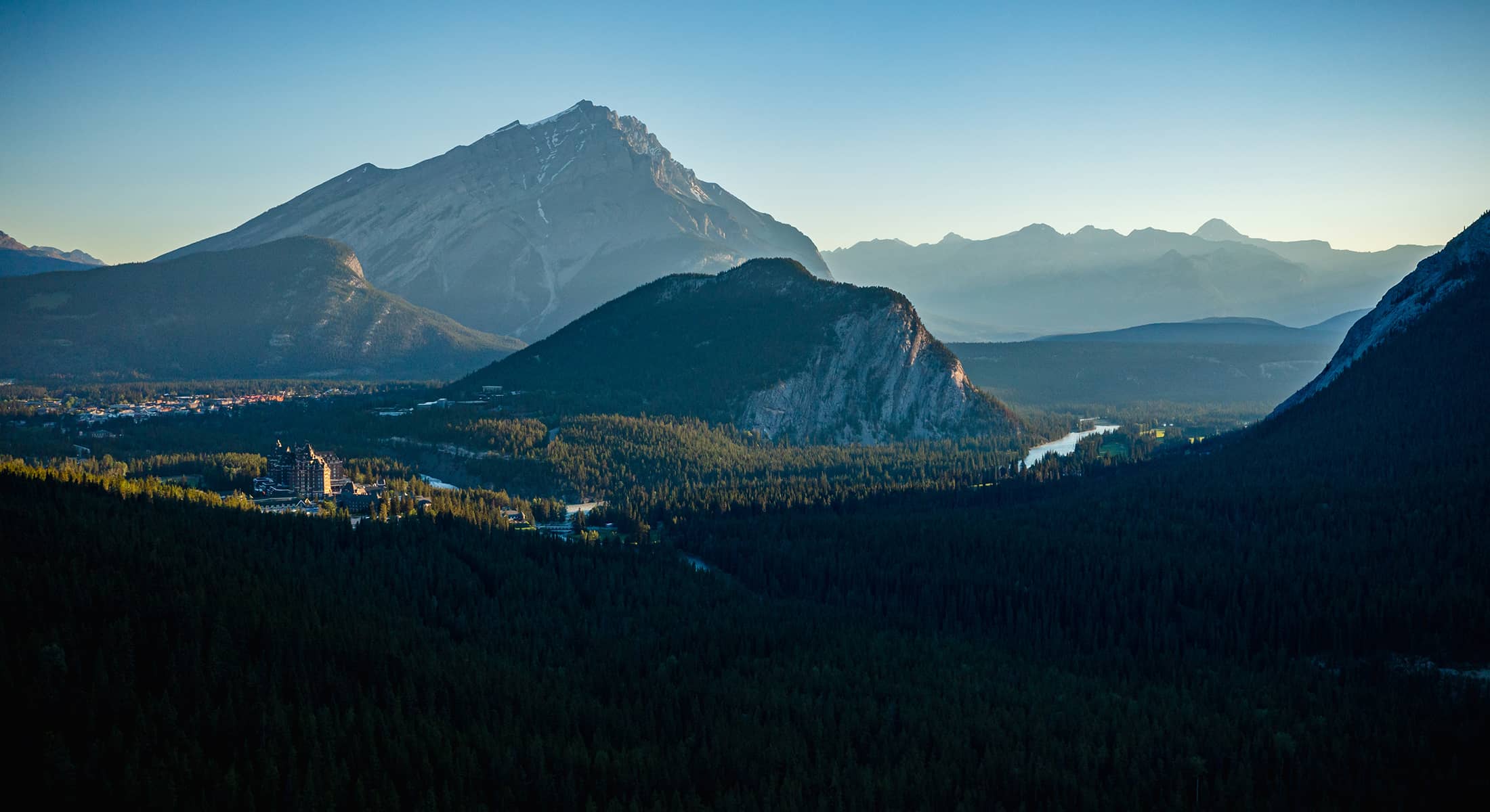 A sweeping view of Banff at sunrise, featuring Cascade Mountain towering behind Tunnel Mountain, with the Bow River valley illuminated by soft morning light.