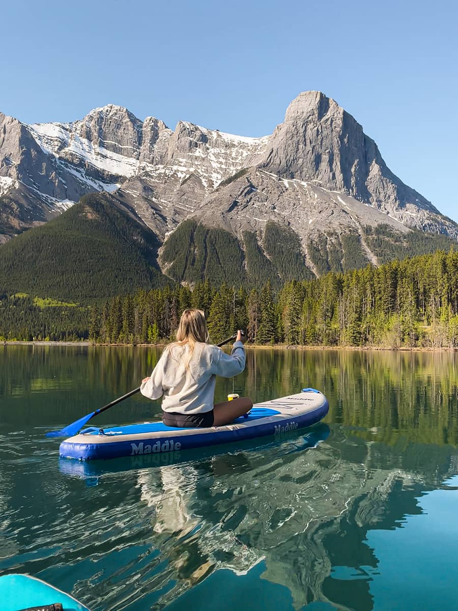 A woman paddles across a calm mountain lake on a sunny day, surrounded by evergreen forests and towering snow-capped peaks reflected in the water.