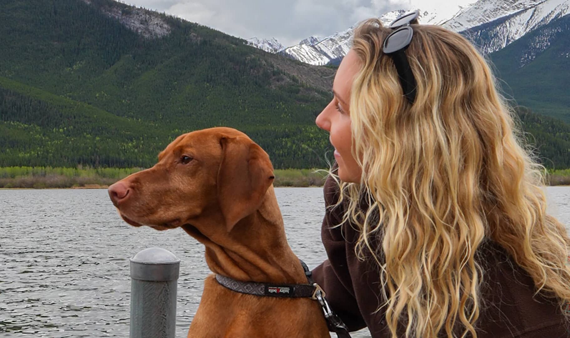 A woman and her dog stand together at Vermillion Lakes in Banff, gazing across the calm water toward the surrounding mountain peaks.