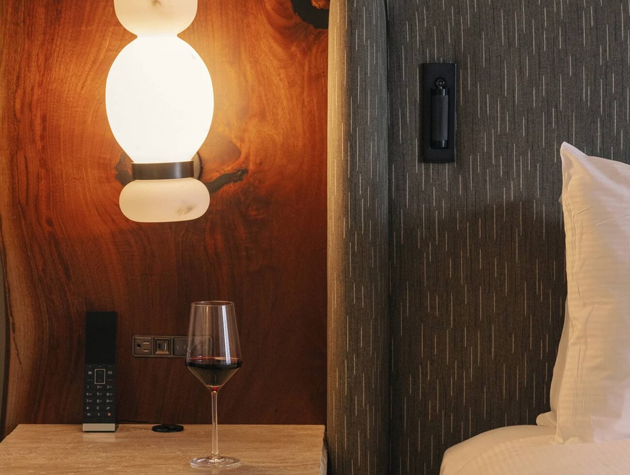 A cozy Rimrock Banff guest room detail featuring a softly lit bedside lamp, textured headboard, and a glass of red wine resting on a stone nightstand.