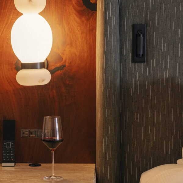 en:: A cozy Rimrock Banff guest room detail featuring a softly lit bedside lamp, textured headboard, and a glass of red wine resting on a stone nightstand. | fr:: Une chambre Rimrock Banff confortable avec une lampe de chevet à l’éclairage subtil, une tête de lit texturée et un verre de vin rouge reposant sur une table de nuit en pierre. | es:: Un detalle de una acogedora habitación de Rimrock Banff que cuenta con una lámpara de noche que ilumina suavemente, cabecera texturizada y una copa de vino tinto sobre una mesilla de piedra. |