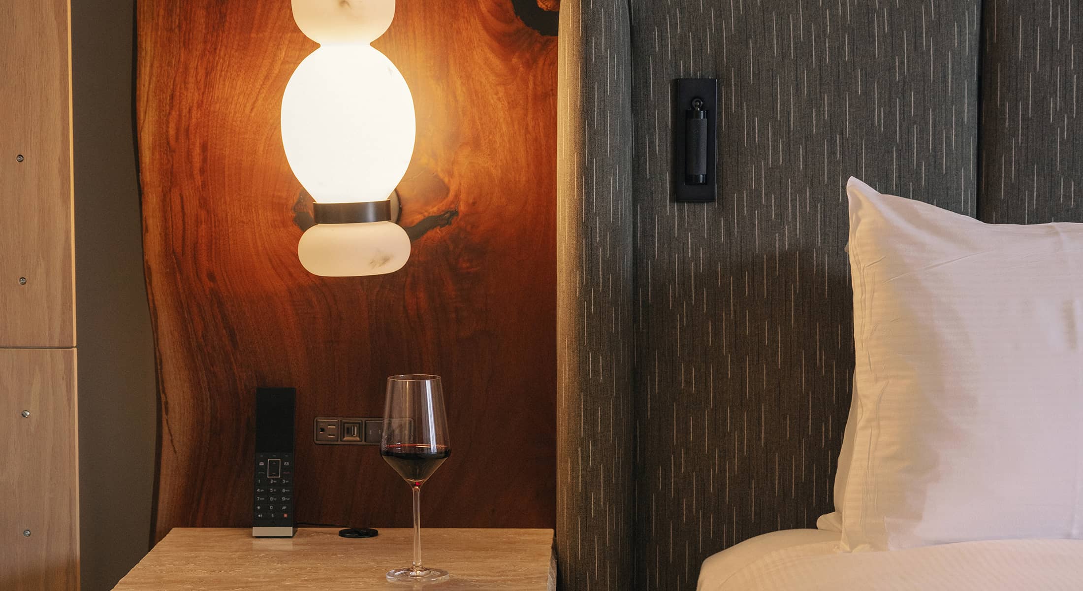 A cozy Rimrock Banff guest room detail featuring a softly lit bedside lamp, textured headboard, and a glass of red wine resting on a stone nightstand.