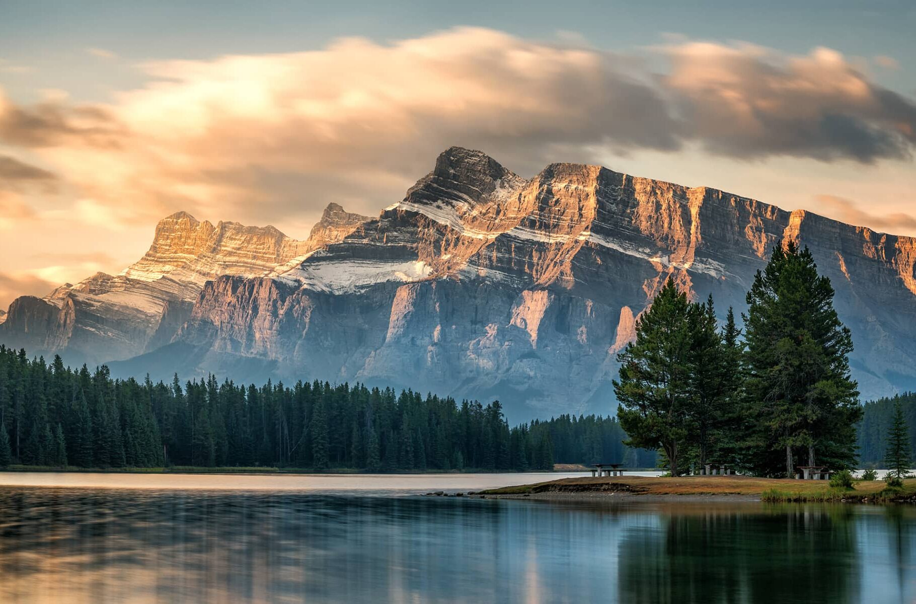 Rundle Mountain reflected in the calm waters of Cascade Ponds, surrounded by evergreens and dawn alpine sky.