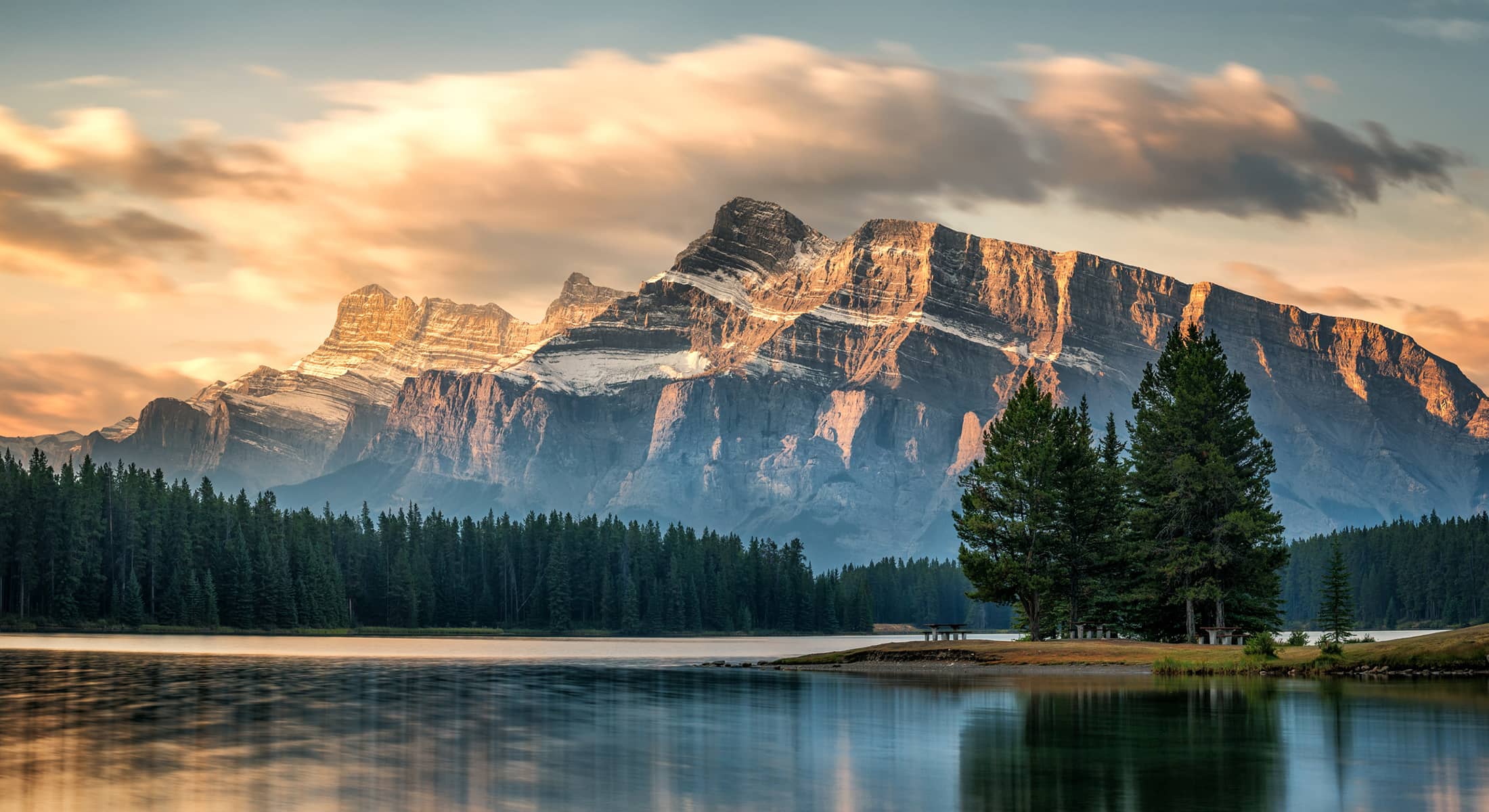 Rundle Mountain reflected in the calm waters of Cascade Ponds, surrounded by evergreens and dawn alpine sky.