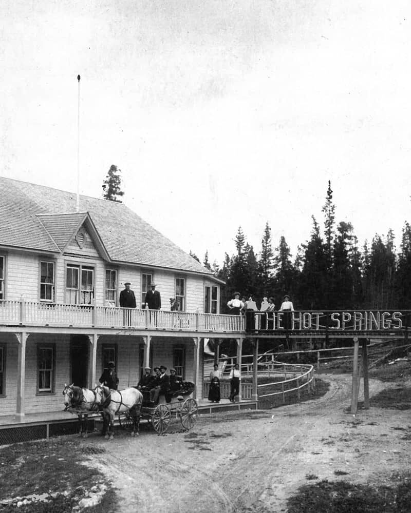 A historic black-and-white photo of the Hot Springs Hydro Hotel in Banff, with guests on the porch and a horse-drawn carriage outside surrounded by forest.