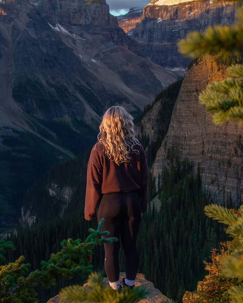 Person standing on a rocky viewpoint surrounded by evergreen trees during golden hour on the summit of Little Beehive in Lake Louise, overlooking dramatic mountain peaks and forested valleys.