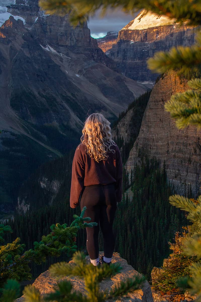 Person standing on a rocky viewpoint surrounded by evergreen trees during golden hour on the summit of Little Beehive in Lake Louise, overlooking dramatic mountain peaks and forested valleys.