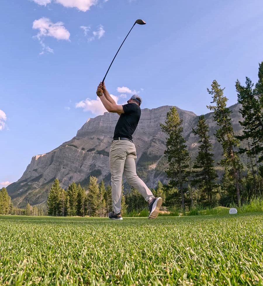 A golfer follows through on a powerful drive at Banff Springs Golf Course surrounded by dramatic mountain peaks under a bright blue sky.