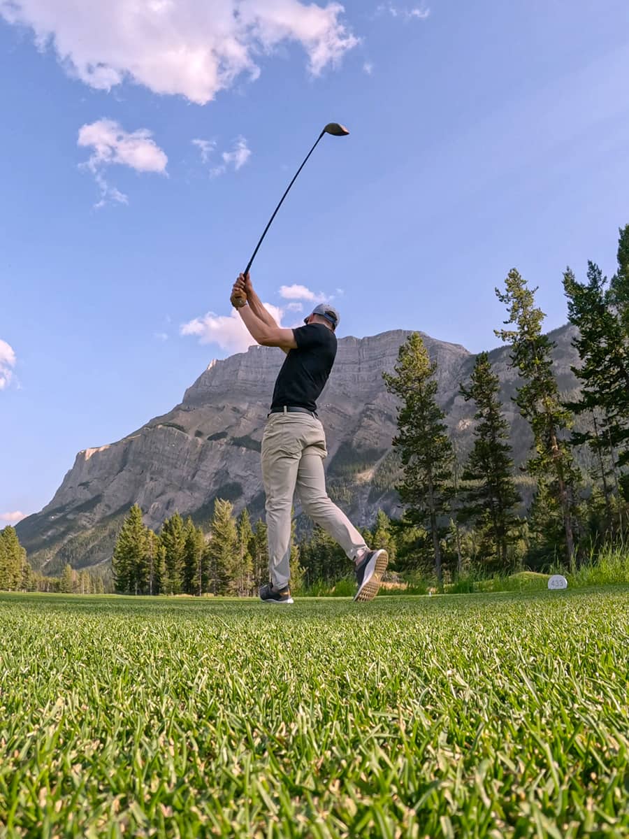 A golfer follows through on a powerful drive at Banff Springs Golf Course surrounded by dramatic mountain peaks under a bright blue sky.