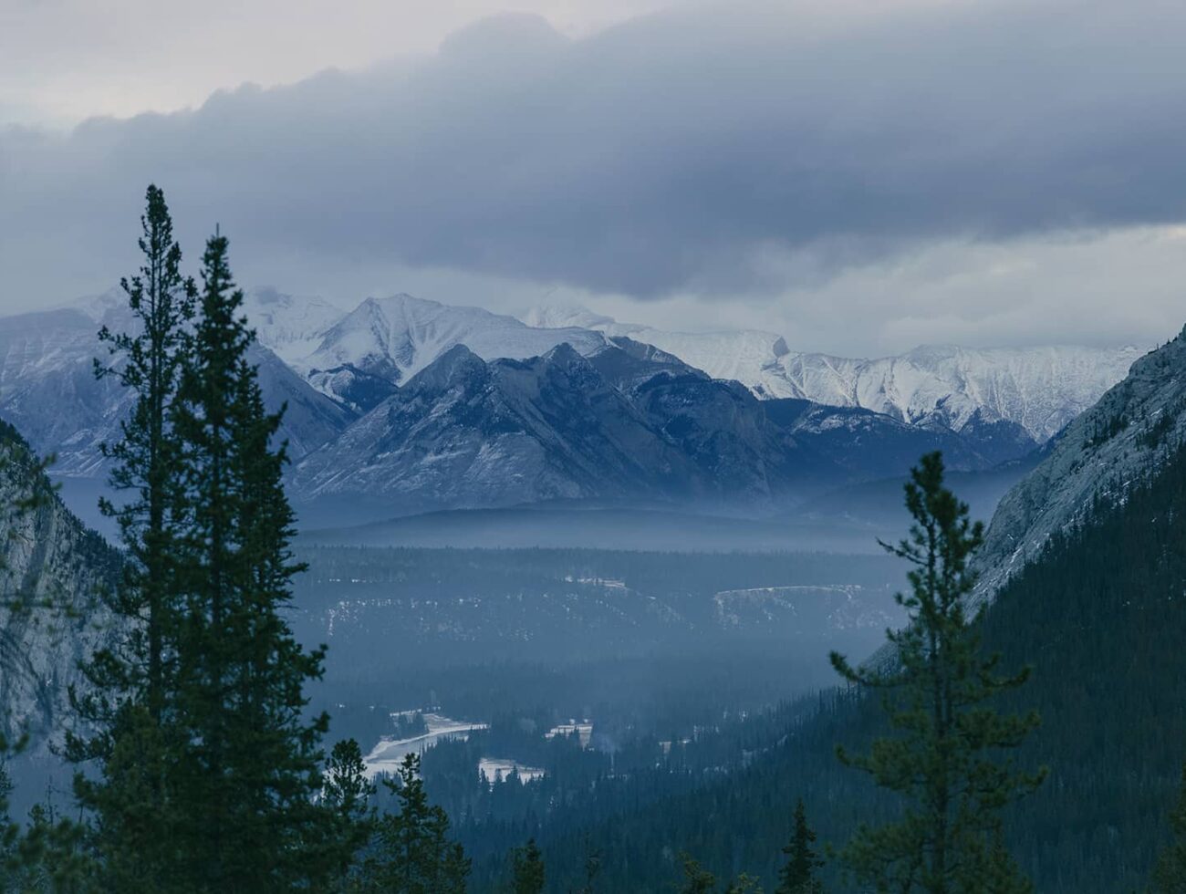 Snow-dusted peaks of the Canadian Rockies rise beyond a misty valley and dense evergreen forest under moody, overcast skies near Banff National Park.