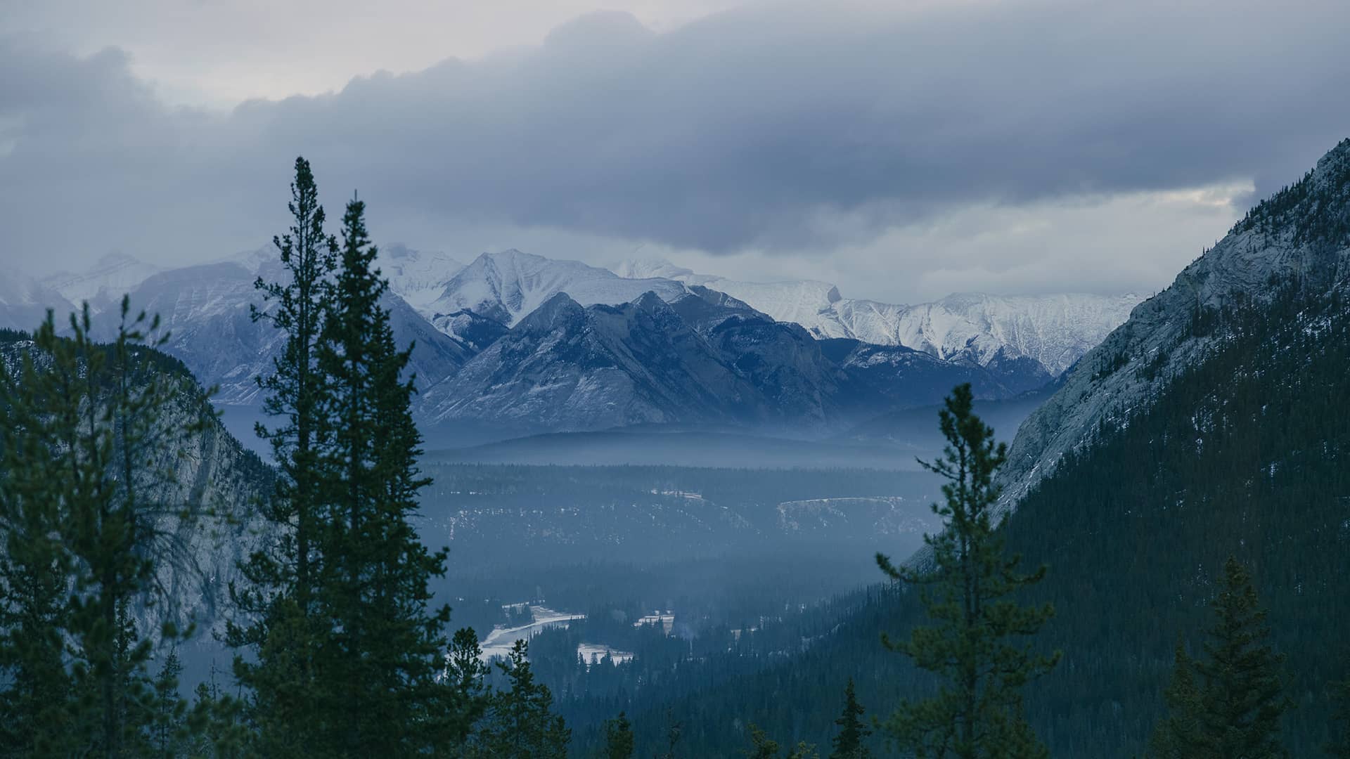Snow-dusted peaks of the Canadian Rockies rise beyond a misty valley and dense evergreen forest under moody, overcast skies near Banff National Park.
