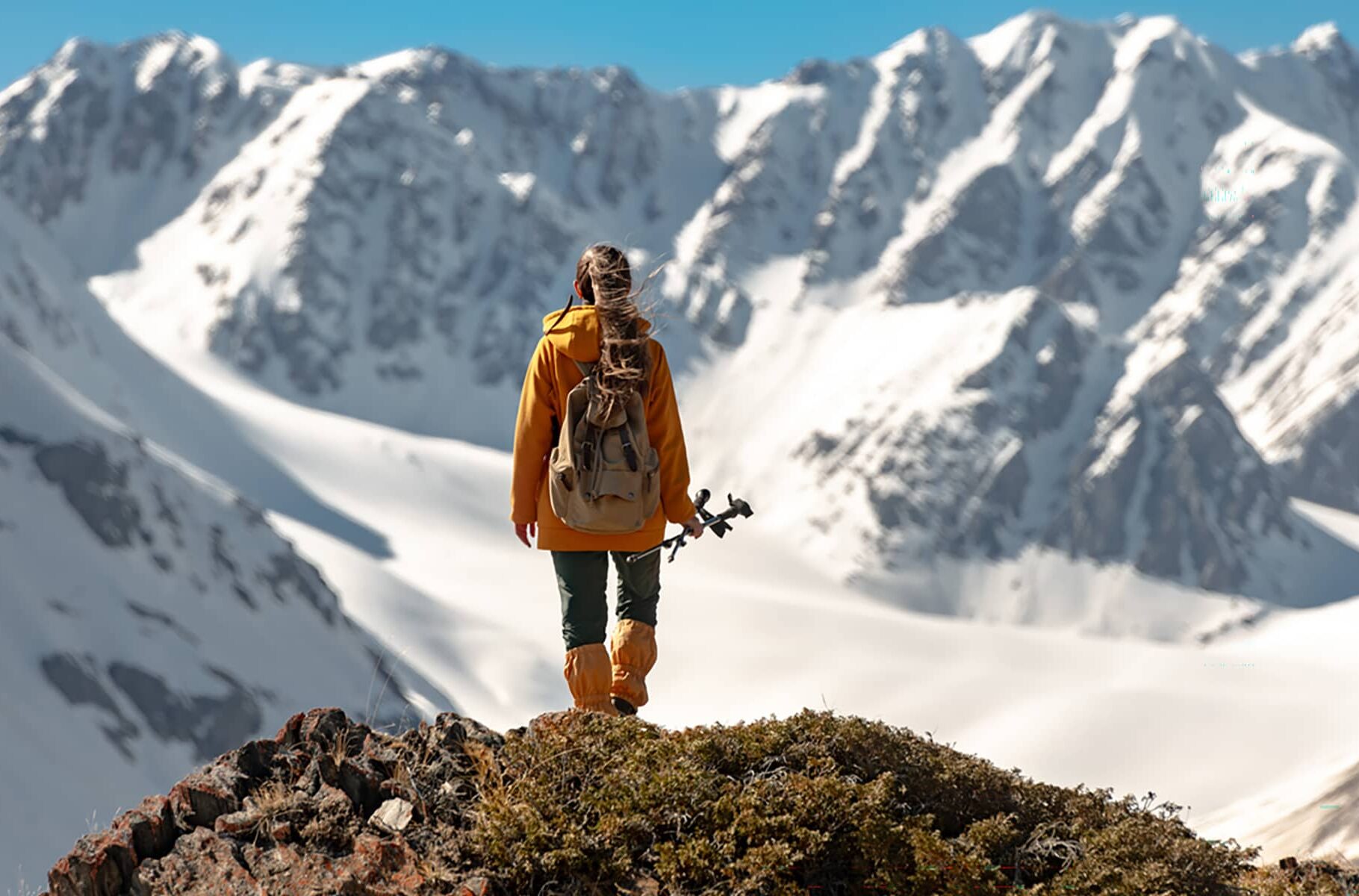 Hiker standing on a rocky ridge, looking out across snow-covered alpine peaks under a clear blue sky.