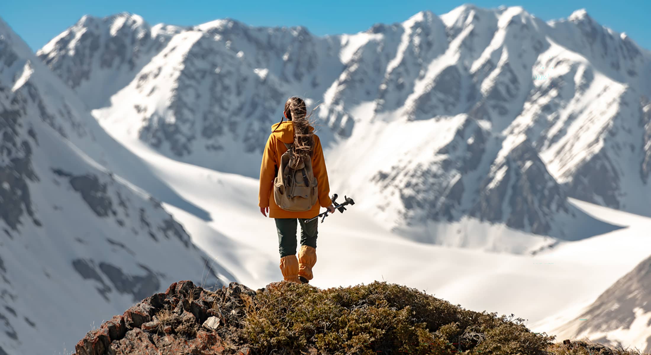 Hiker standing on a rocky ridge, looking out across snow-covered alpine peaks under a clear blue sky.