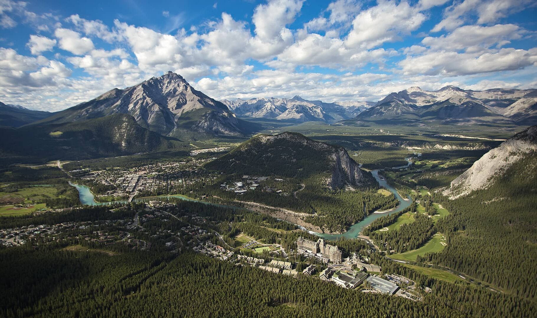 A sweeping aerial view of the Town of Banff surrounded by winding rivers, deep forests, and towering Rocky Mountain peaks under a bright blue sky.