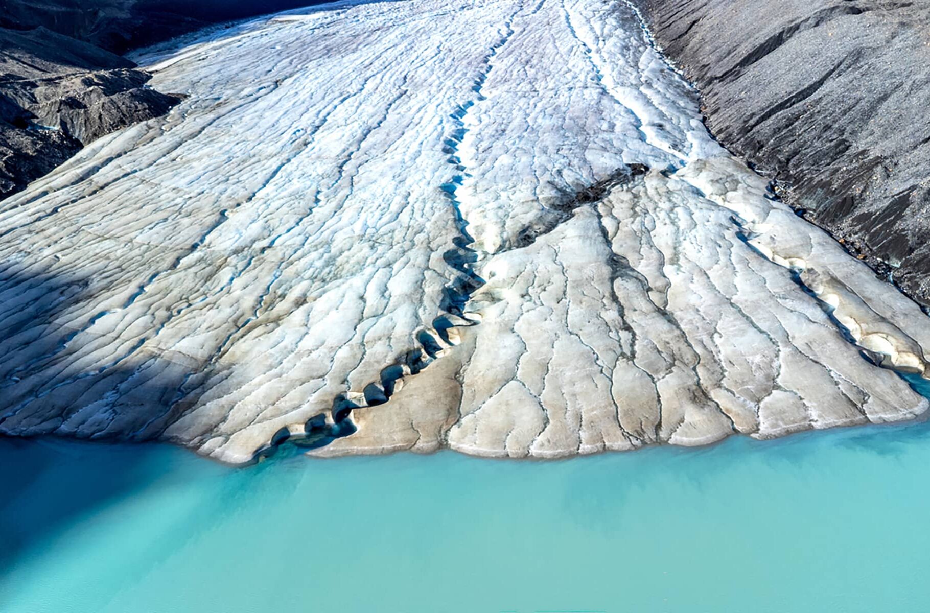 Aerial view of the Athabasca Glacier’s textured ice edge meeting turquoise meltwater, showing crevasses and flowing ice patterns.