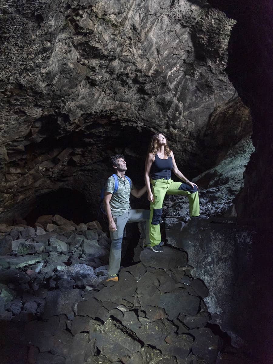 Two people exploring a cave with headlamps, standing on uneven rock formations beneath a high stone ceiling in Banff National Park.