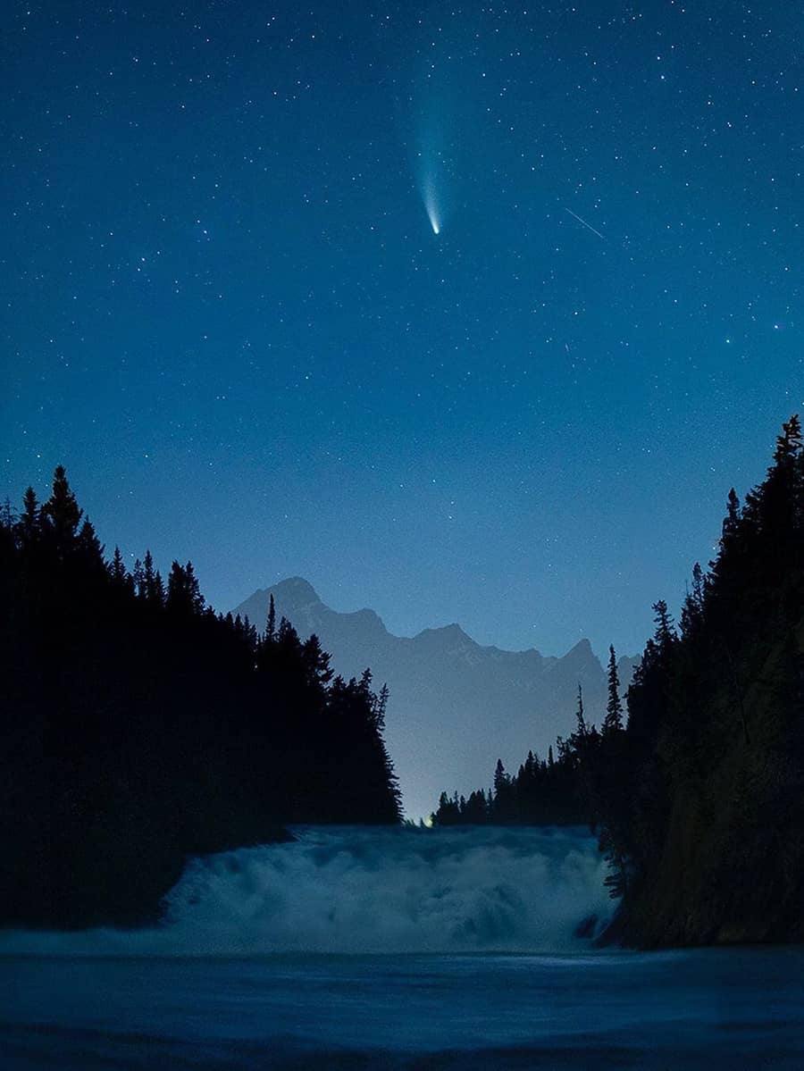 Bow Falls at night with rushing water framed by dark pines, a glowing comet streaking across a clear star-filled sky, and mountain silhouettes beyond.