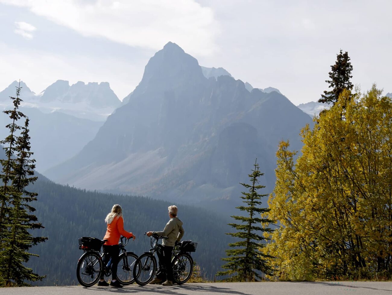 Distant view of mature couple pausing with e-bicycles on Moraine Lake road in autumn, mountain peaks and golden leafed trees in distance, Banff National Park, Canadian Rockies