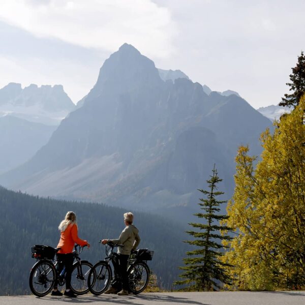 Distant view of mature couple pausing with e-bicycles on Moraine Lake road in autumn, mountain peaks and golden leafed trees in distance, Banff National Park, Canadian Rockies