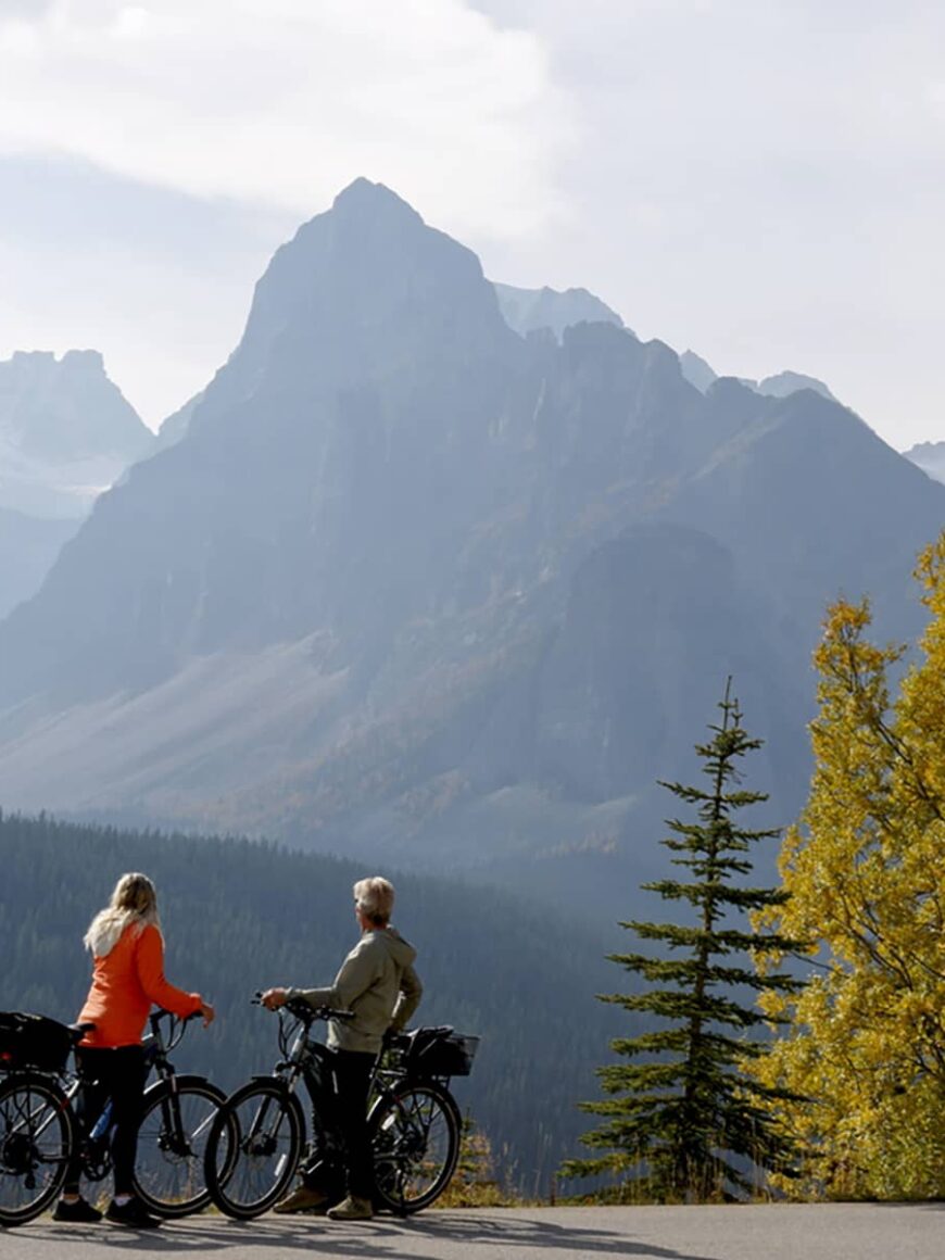 Distant view of mature couple pausing with e-bicycles on Moraine Lake road in autumn, mountain peaks and golden leafed trees in distance, Banff National Park, Canadian Rockies