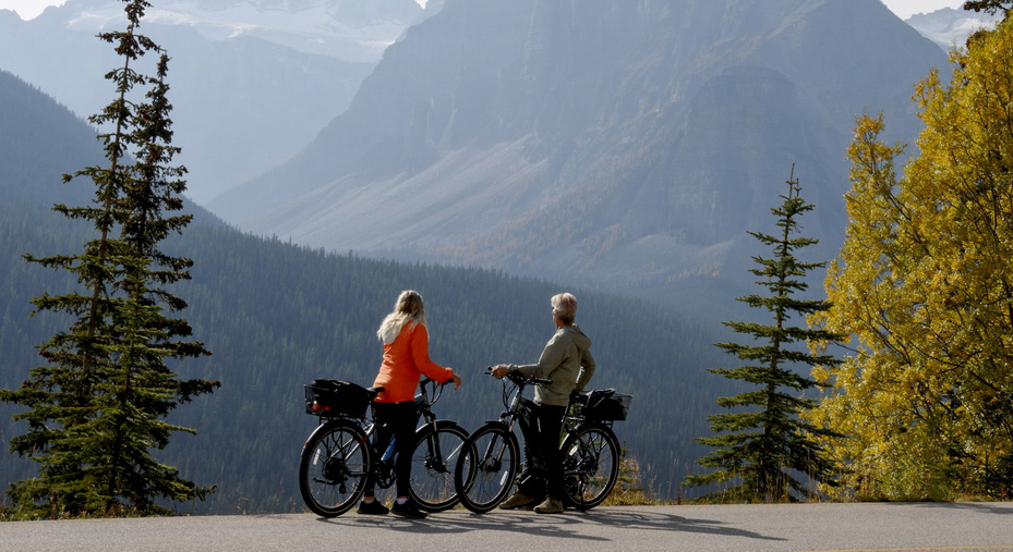 Distant view of mature couple pausing with e-bicycles on Moraine Lake road in autumn, mountain peaks and golden leafed trees in distance, Banff National Park, Canadian Rockies