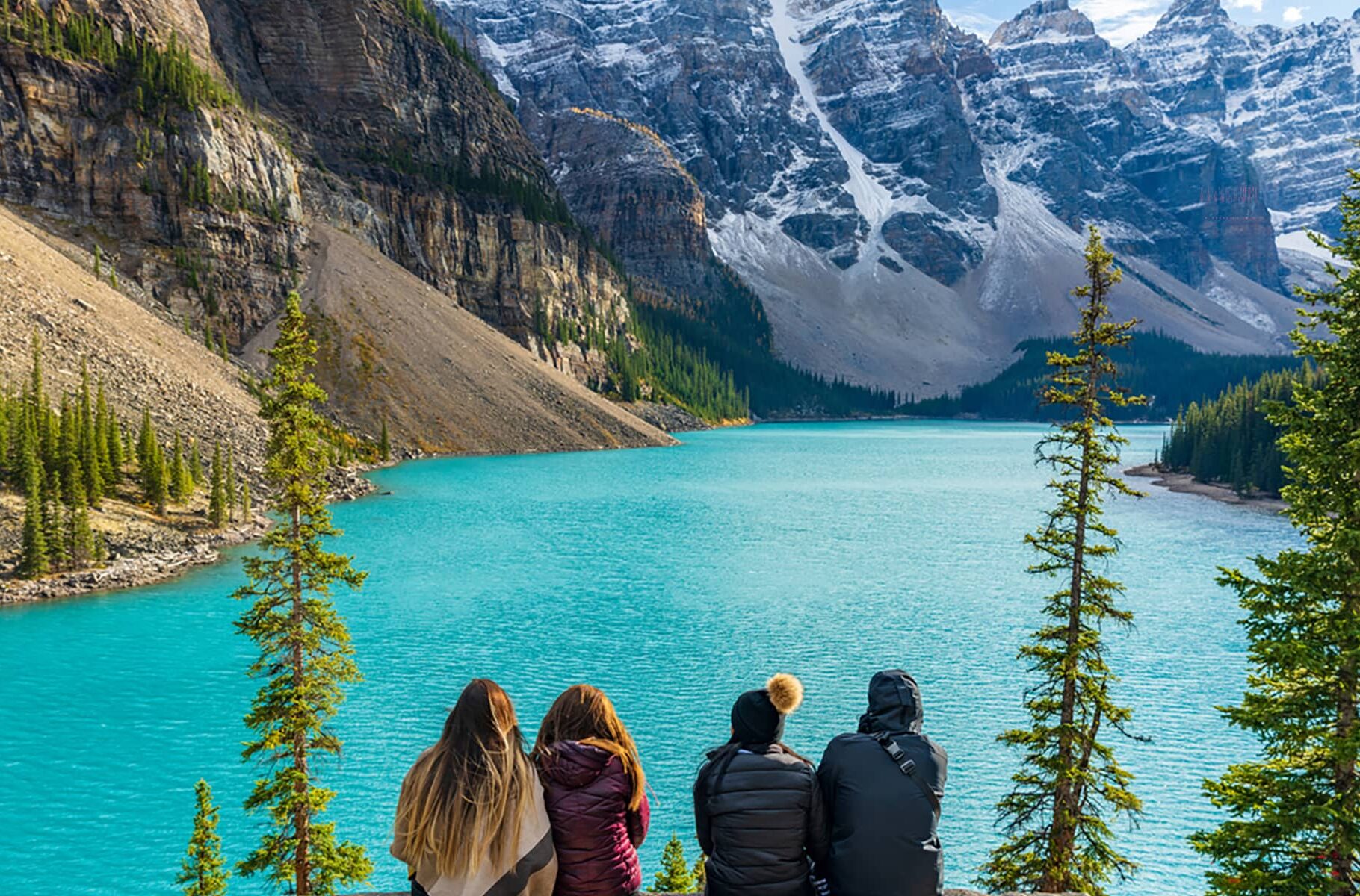 Four young women on vacation, sitting together by the turquoise Moraine Lake, admiring the snow-capped Ten Peaks. Banff National Park, Alberta, Canada.