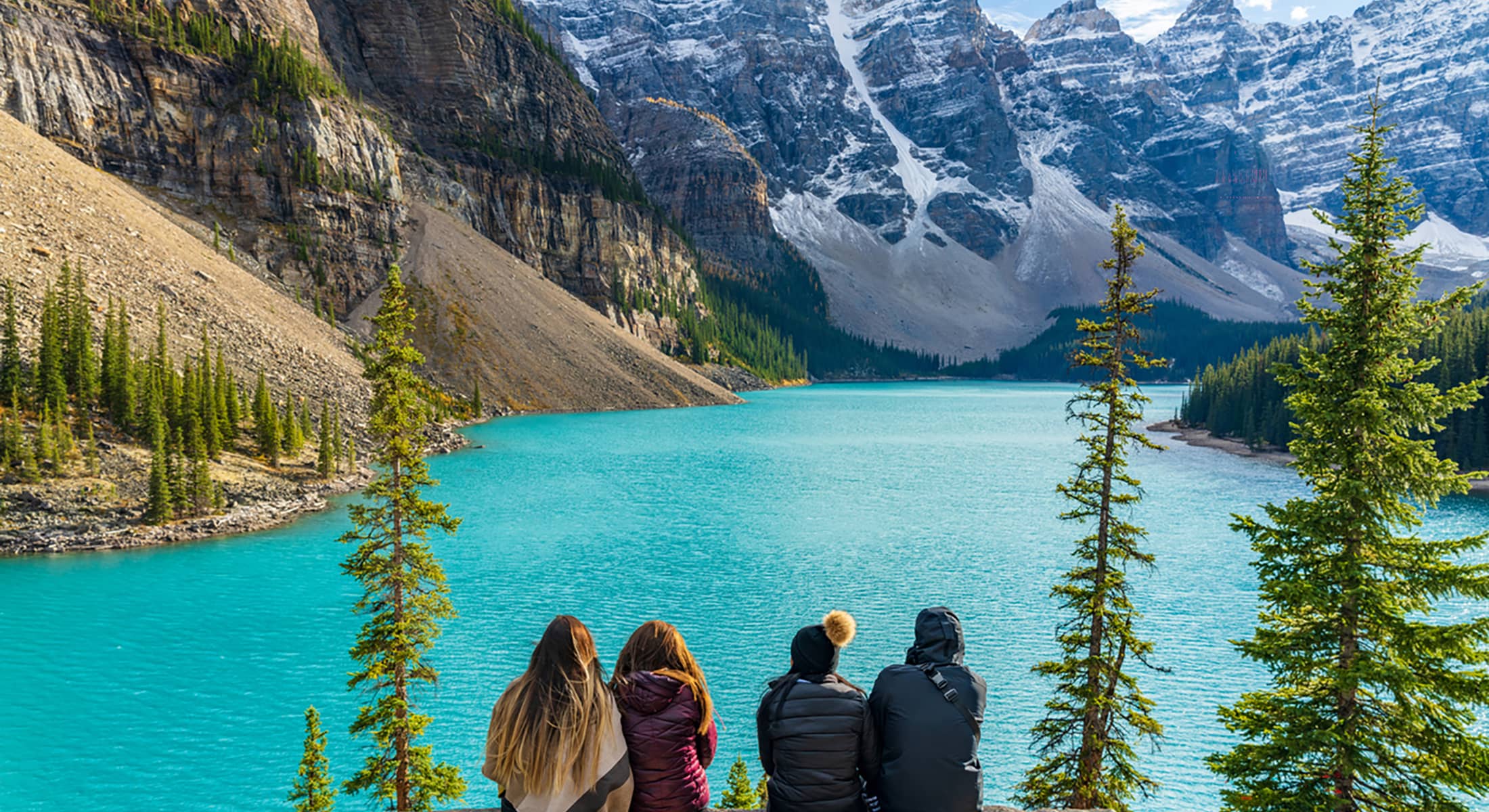 Four young women on vacation, sitting together by the turquoise Moraine Lake, admiring the snow-capped Ten Peaks. Banff National Park, Alberta, Canada.