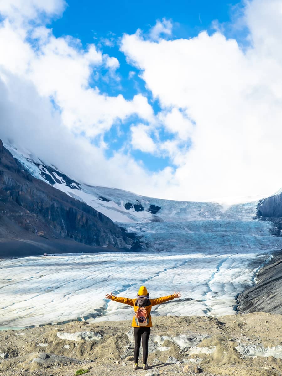 Person standing with arms outstretched at the edge of the Athabasca Glacier, surrounded by ice, rock, and alpine peaks.