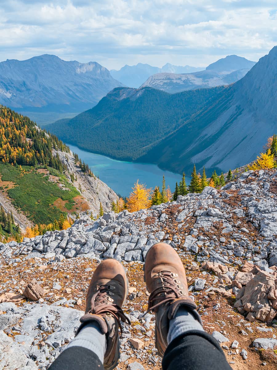 Hiking boots resting on a rocky ledge, overlooking a deep mountain valley, alpine lake, and layered peaks beyond.