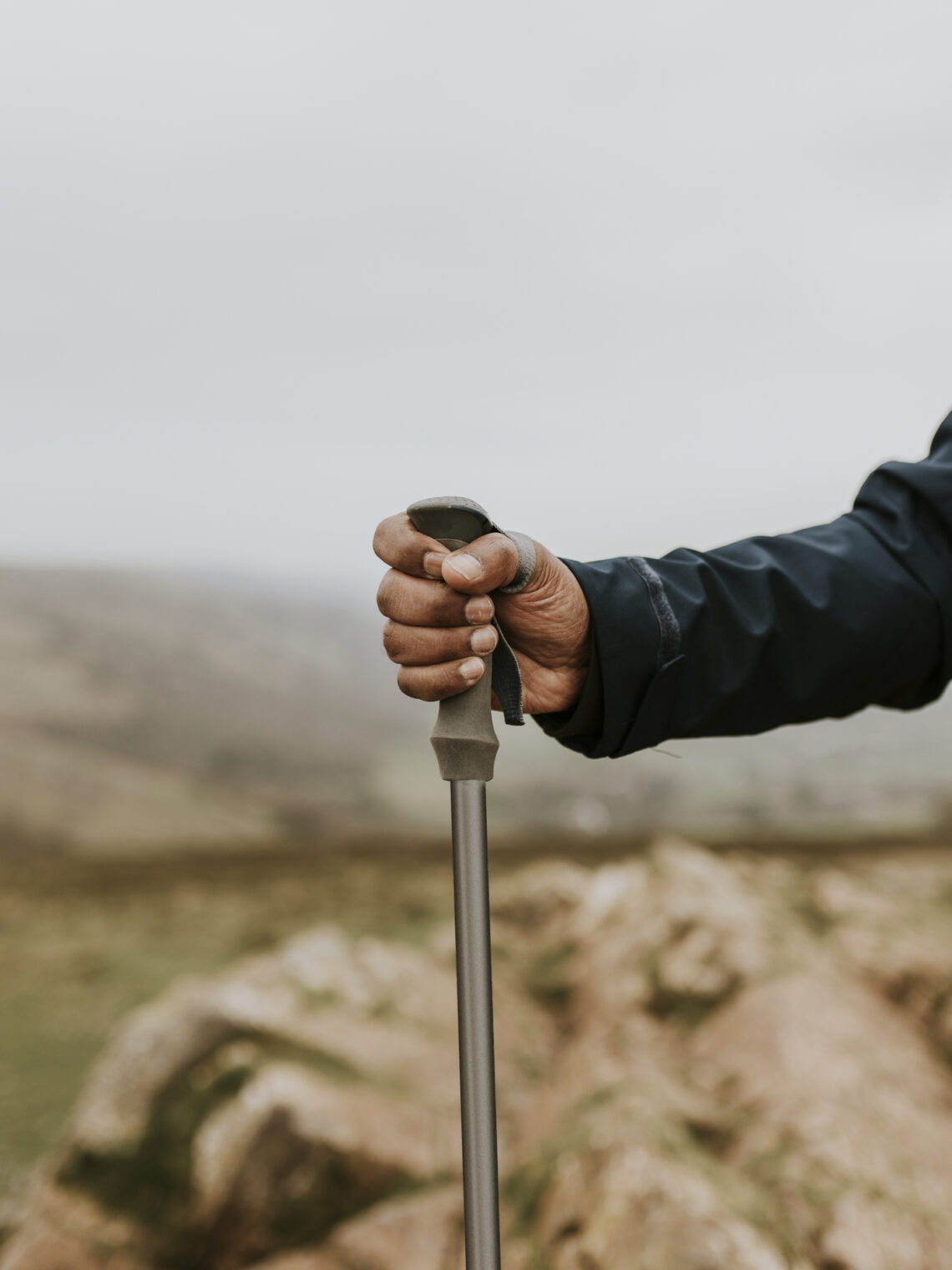 A close-up of a hiker’s hand gripping a trekking pole, with soft, hazy mountain terrain stretching out in the distance.