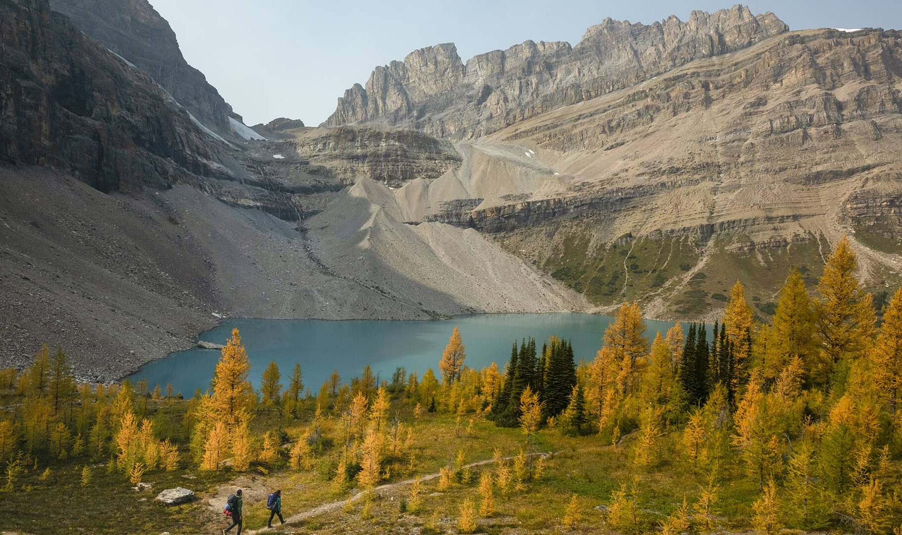 Two hikers walk through golden larch trees toward a turquoise alpine lake, framed by rugged cliffs and dramatic late-season mountain scenery.