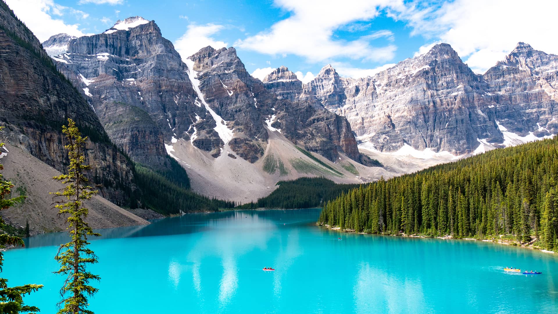 Turquoise waters of Moraine Lake framed by pine forest and dramatic rocky peaks beneath a bright alpine sky.