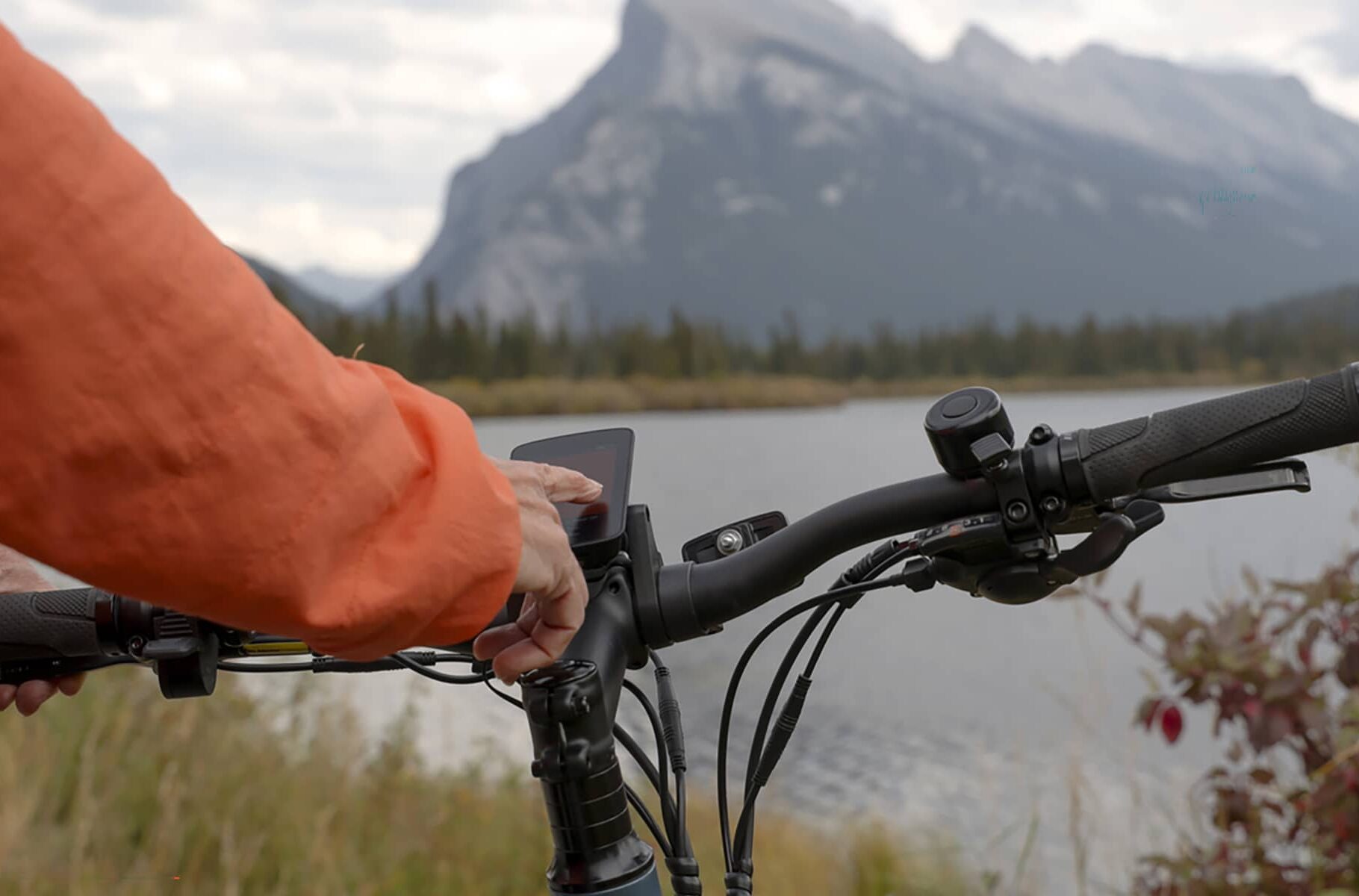Detail of finger touching digital screen on e-bicycle by lakeshore, Vermillion Lakes, Mount Rundle in distance, Banff National Park, Canadian Rockies