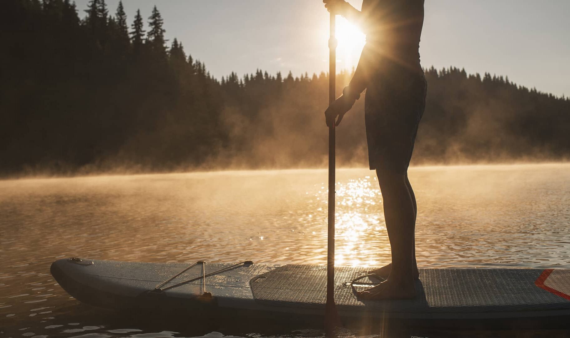A person paddleboarding on a calm mountain lake at sunrise, with golden light, rising mist, and forested shoreline in the background.