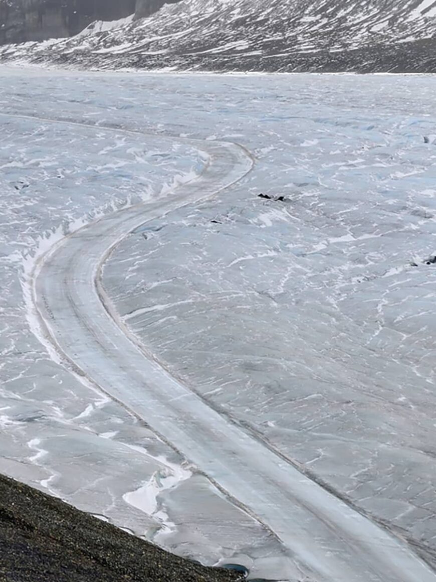 View of the Athabasca Glacier showing a winding track across textured ice, with surrounding rock and alpine slopes.