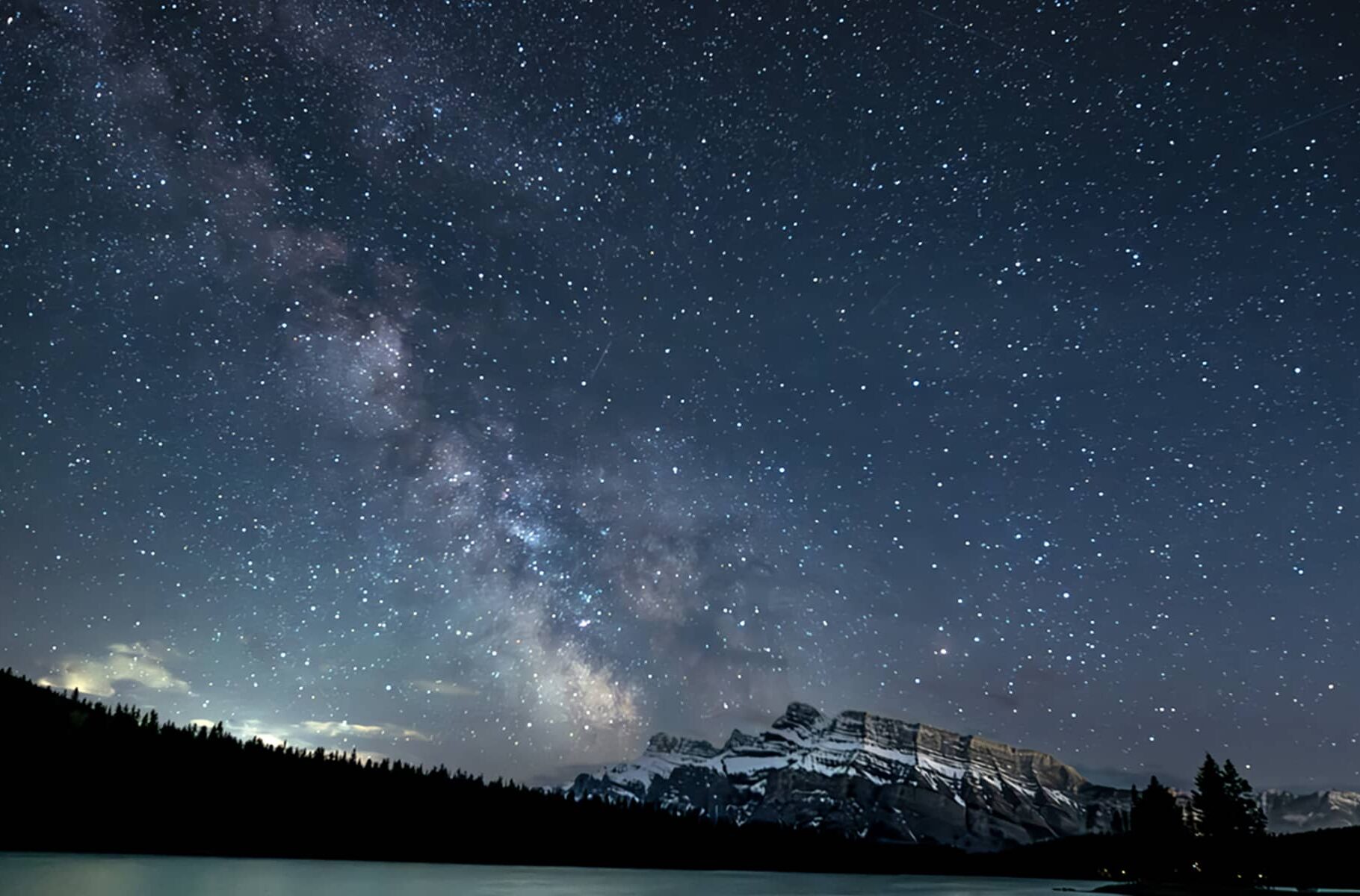 The Milky Way visible above Mount Rundle, reflected in calm water and framed by forested shoreline.