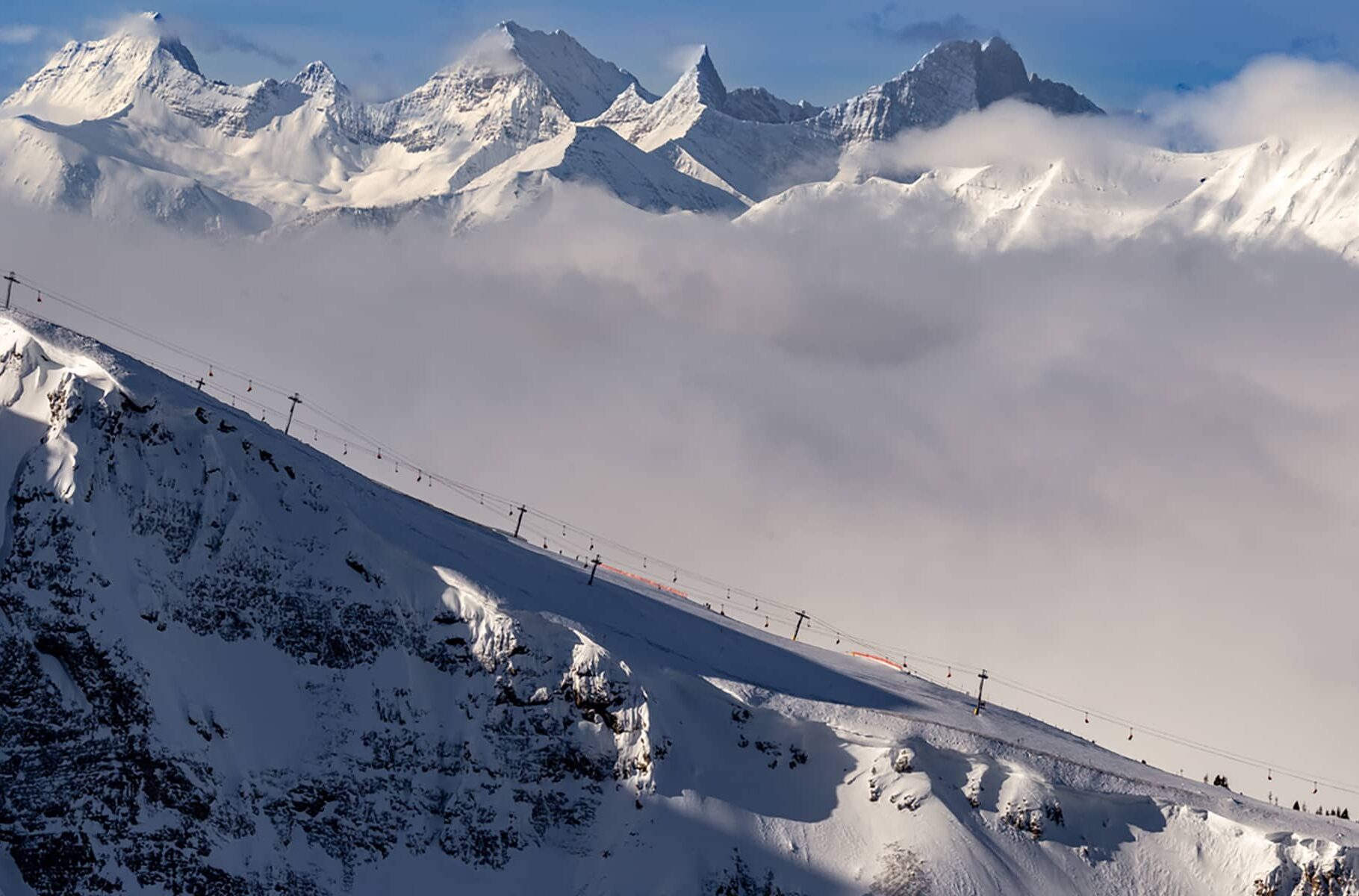 A high-alpine view at Sunshine Village showing chairlifts crossing a snow-covered ridge above a low cloud layer and distant mountain peaks.
