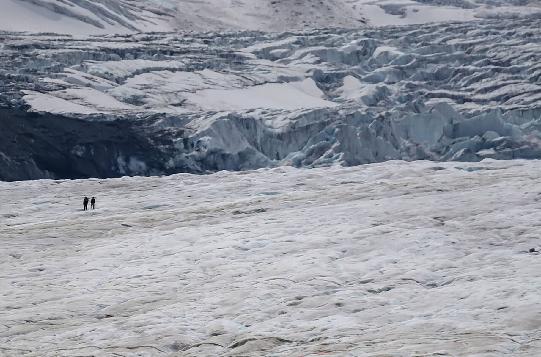 Two people walking across the Athabasca Glacier, set against layered ice formations and surrounding alpine terrain.