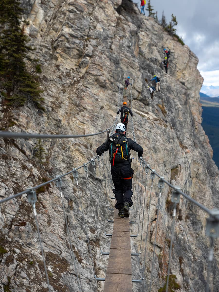A man crosses a narrow suspension bridge on Mount Norquay’s Via Ferrata, harnessed above a dramatic cliffside route.