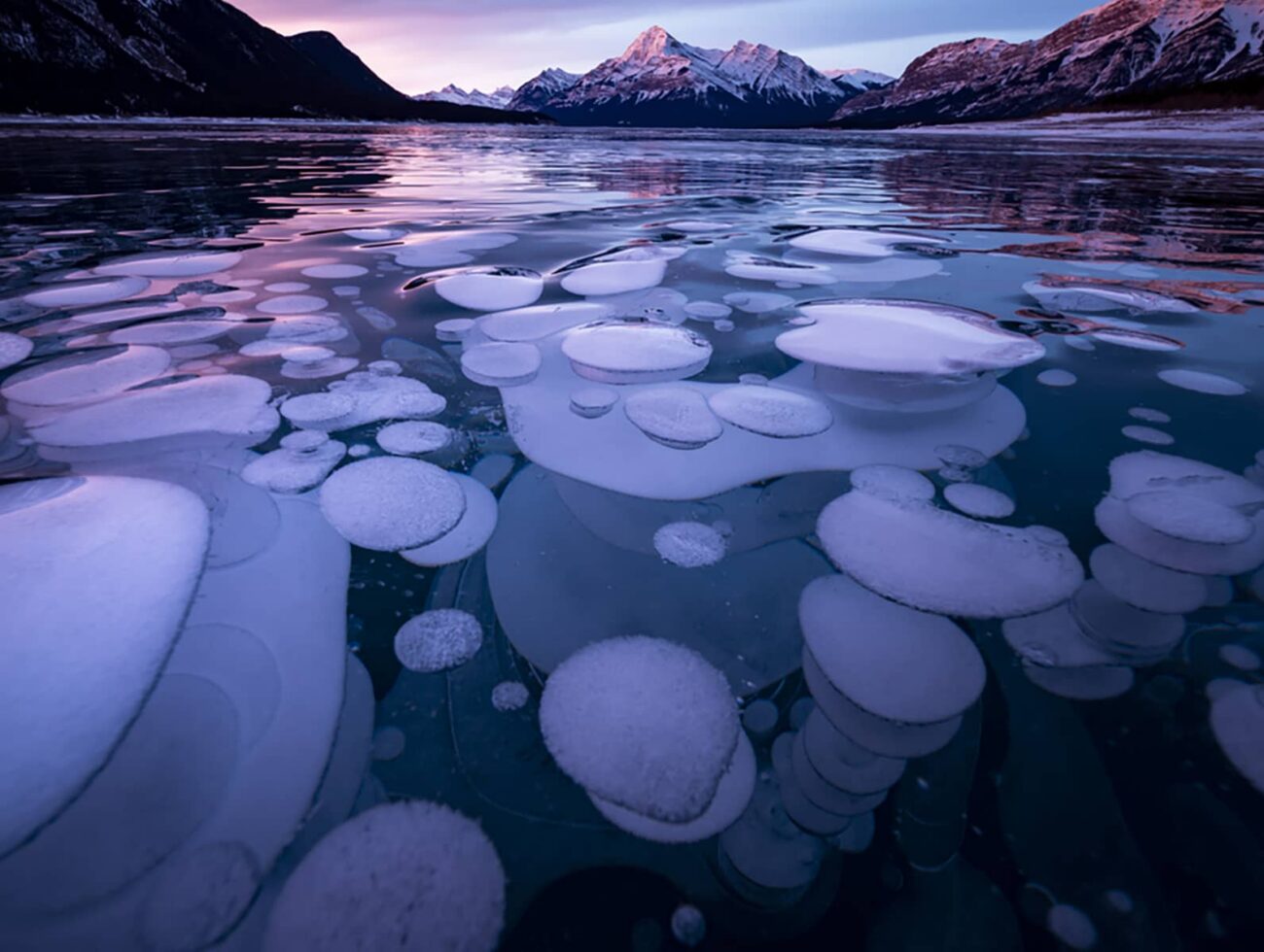 Frozen Abraham Lake at sunset, showing ice bubbles beneath the surface and snow-covered peaks under colorful winter skies.