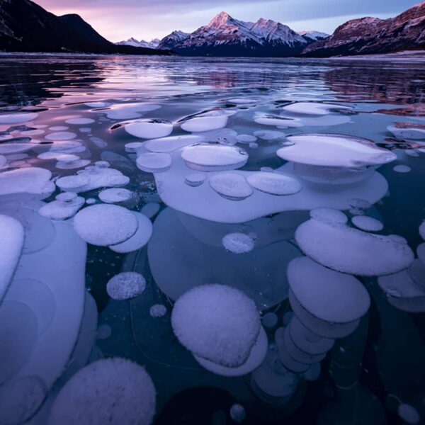 Frozen Abraham Lake at sunset, showing ice bubbles beneath the surface and snow-covered peaks under colorful winter skies.