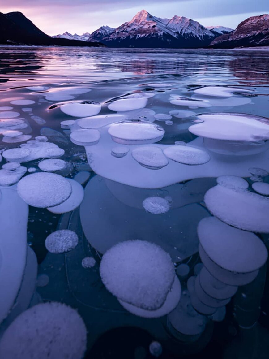 Frozen Abraham Lake at sunset, showing ice bubbles beneath the surface and snow-covered peaks under colorful winter skies.