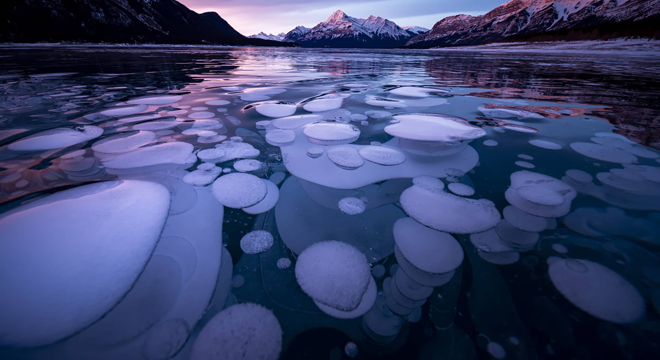Frozen Abraham Lake at sunset, showing ice bubbles beneath the surface and snow-covered peaks under colorful winter skies.