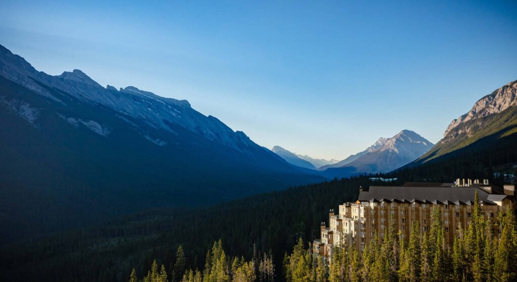 Aerial view of Rimrock Banff perched along Sulphur Mountain, surrounded by dense evergreen forest with sweeping views of the Bow Valley and Rocky Mountain peaks under a clear blue sky.