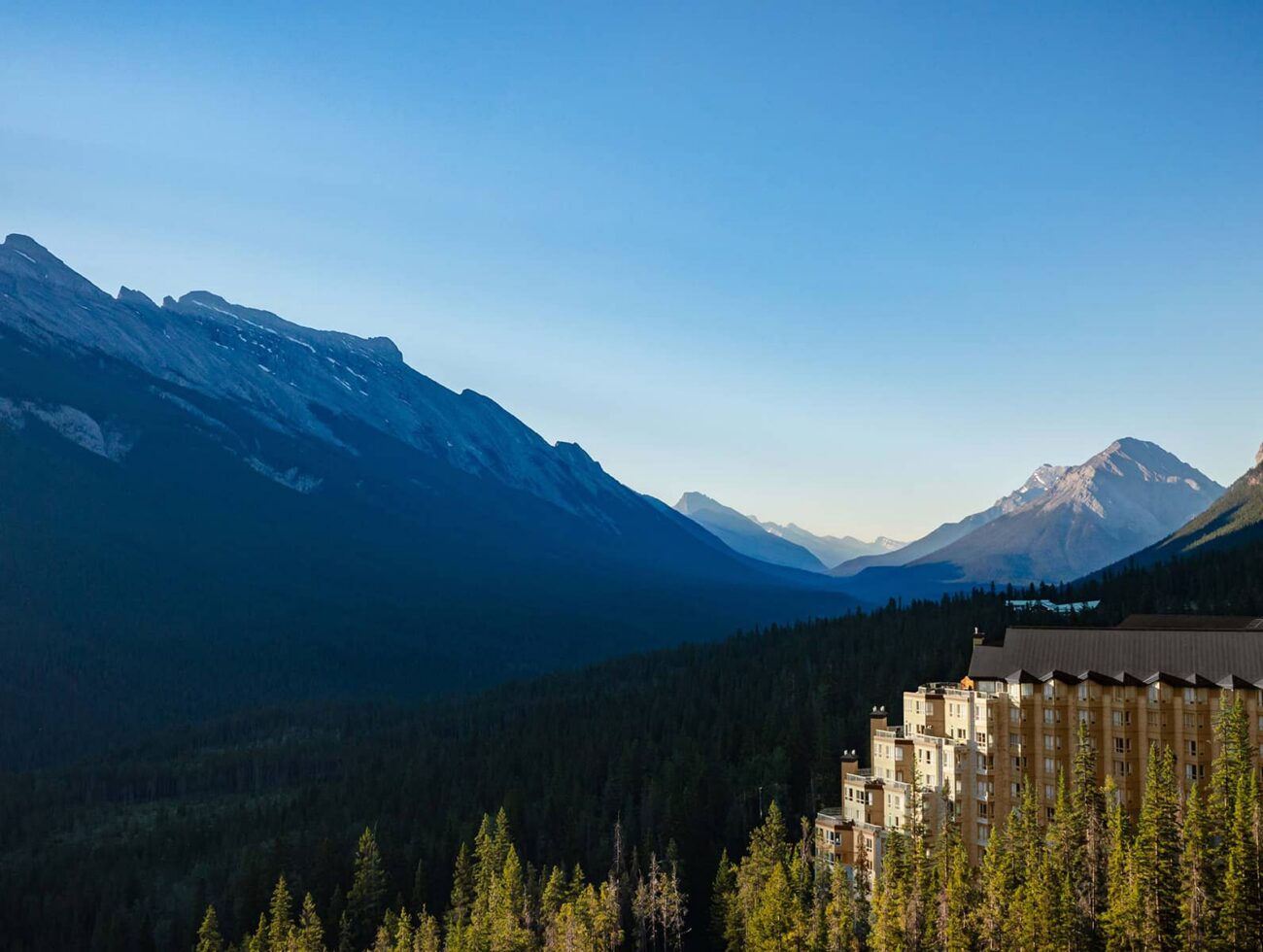 Aerial view of Rimrock Banff perched along Sulphur Mountain, surrounded by dense evergreen forest with sweeping views of the Bow Valley and Rocky Mountain peaks under a clear blue sky.
