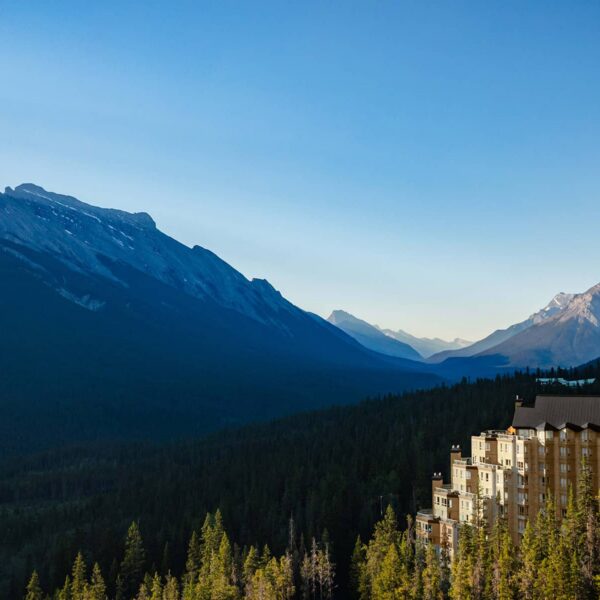 Aerial view of Rimrock Banff perched along Sulphur Mountain, surrounded by dense evergreen forest with sweeping views of the Bow Valley and Rocky Mountain peaks under a clear blue sky.