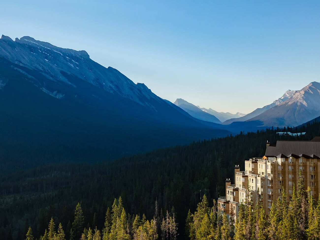 Aerial view of Rimrock Banff perched along Sulphur Mountain, surrounded by dense evergreen forest with sweeping views of the Bow Valley and Rocky Mountain peaks under a clear blue sky.