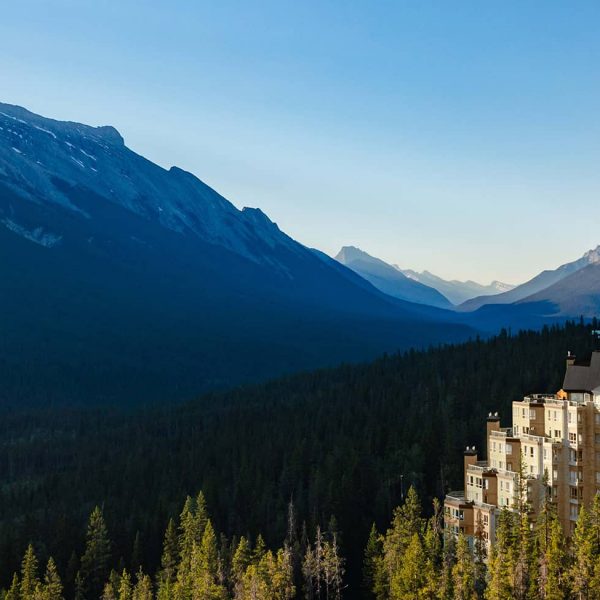 Aerial view of Rimrock Banff perched along Sulphur Mountain, surrounded by dense evergreen forest with sweeping views of the Bow Valley and Rocky Mountain peaks under a clear blue sky.