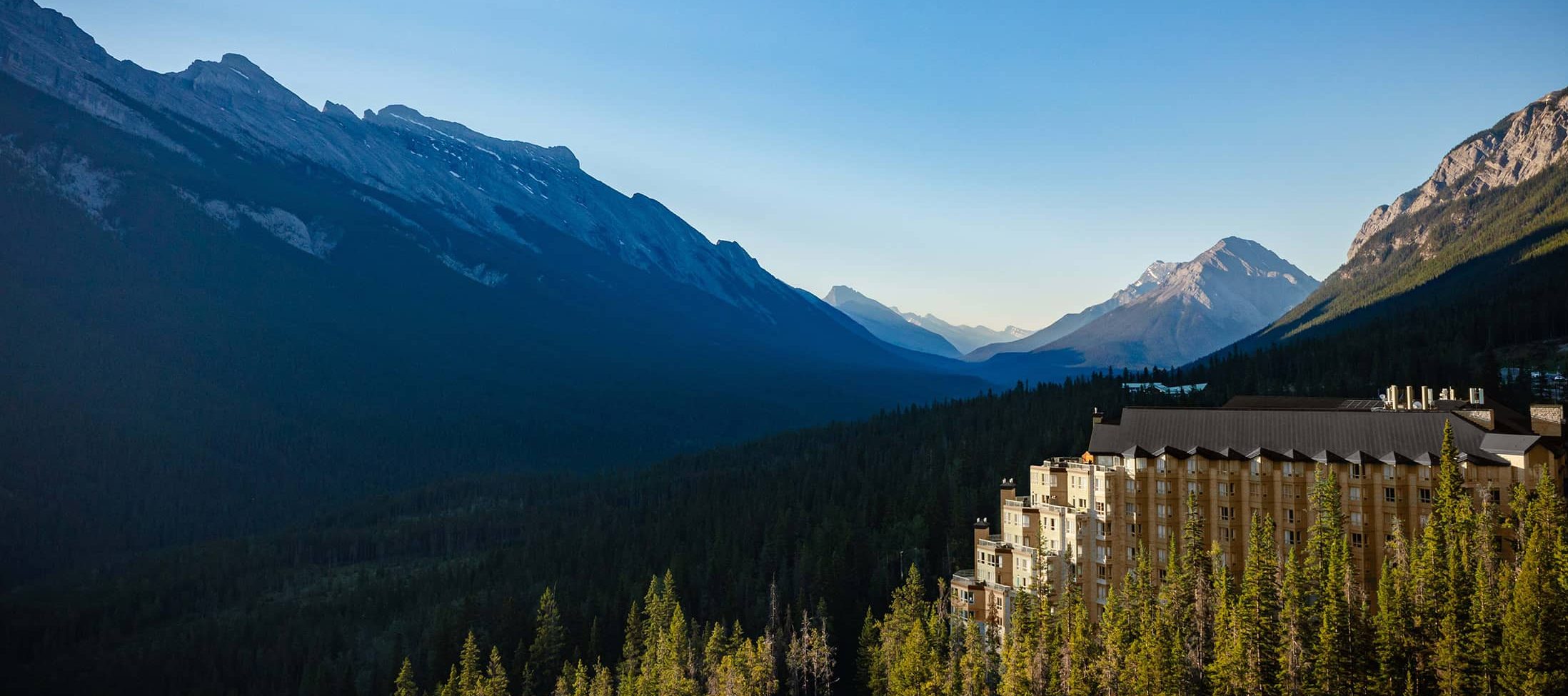 Aerial view of Rimrock Banff perched along Sulphur Mountain, surrounded by dense evergreen forest with sweeping views of the Bow Valley and Rocky Mountain peaks under a clear blue sky.