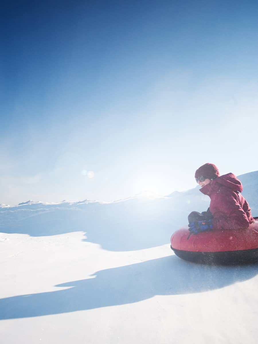 A person snow tubing downhill on a snowy slope with mountain views in the distance.